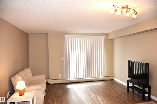 Spacious living area featuring wood-finish flooring, neutral wall tones, and a sliding glass door with vertical blinds - 103 920 156 Street Nw, Edmonton, AB - Indoor Photo Showing Other Room