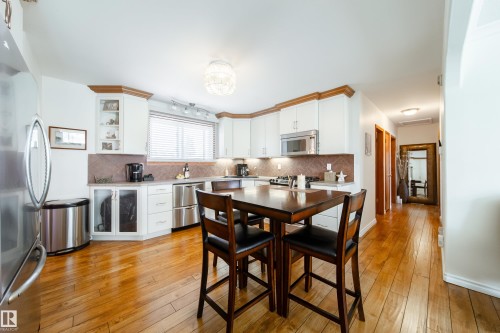Bright kitchen featuring a corner window with horizontal blinds, white cabinetry with wood trim, stainless steel appliances, and wood-finish flooring - 8932 154 Street, Edmonton, AB - Indoor