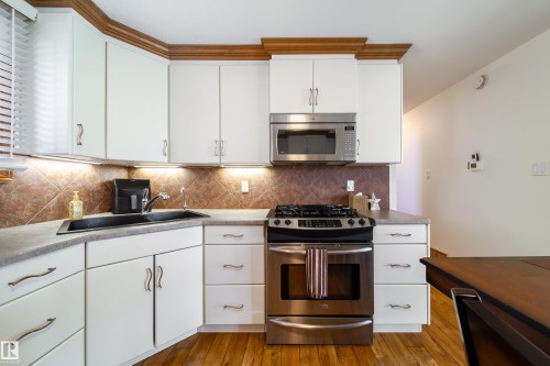 Kitchen featuring white cabinetry with decorative wood crown molding, a stainless steel range with a gas cooktop, a built-in microwave, a black corner sink, and a tiled backsplash - 8932 154 Street, Edmonton, AB - Indoor Photo Showing Kitchen With Double Sink