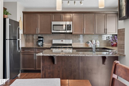 Kitchen featuring rich brown cabinetry, stainless steel appliances, a tile backsplash, and a granite countertop with an undermount sink - 1786 Cunningham Way Sw, Edmonton, AB - Indoor Photo Showing Kitchen With Double Sink With Upgraded Kitchen