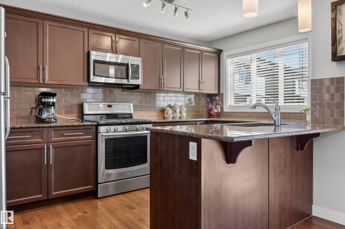 Kitchen featuring wood-finish cabinetry, granite-style countertops, stainless steel appliances, a tiled backsplash, and wood-finish flooring - 1786 Cunningham Way Sw, Edmonton, AB - Indoor Photo Showing Kitchen
