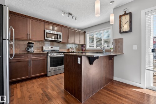 Kitchen featuring wood-finish flooring, dark wood cabinetry, stainless steel appliances, and a central island with an integrated sink - 1786 Cunningham Way Sw, Edmonton, AB - Indoor Photo Showing Kitchen