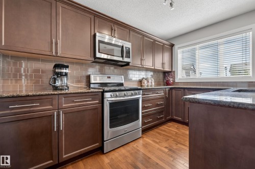 Kitchen featuring rich brown cabinetry, stainless steel appliances, granite countertops, and a tiled backsplash - 1786 Cunningham Way Sw, Edmonton, AB - Indoor Photo Showing Kitchen