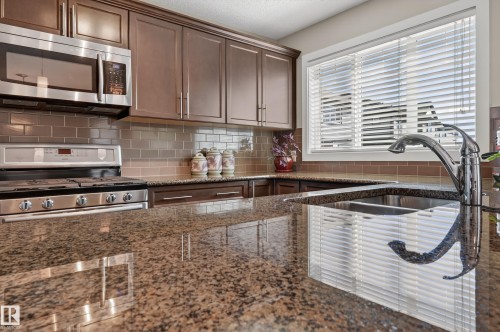 Kitchen featuring extensive stone countertops, a double basin undermount sink with a high-arc faucet, stainless steel appliances, wood cabinetry, and a subway tile backsplash - 1786 Cunningham Way Sw, Edmonton, AB - Indoor Photo Showing Kitchen With Double Sink