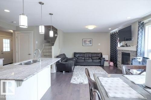 Open-concept living area featuring a kitchen island with dual basin sink and high-arc faucet, wood-finish flooring, a stone-clad fireplace, and a staircase with white railings - 16619 14 Avenue, Edmonton, AB - Indoor Photo Showing Kitchen With Fireplace With Double Sink With Upgraded Kitchen