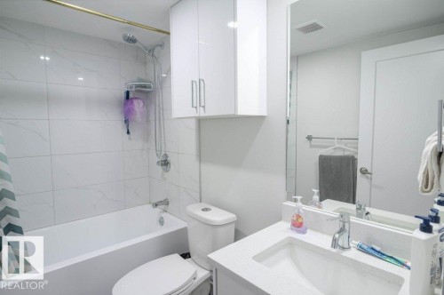 Bathroom featuring a shower-tub combination with white tile surround, a built-in vanity with a white countertop and integrated sink, a wall-mounted mirror, and overhead white cabinetry - 16619 14 Avenue, Edmonton, AB - Indoor Photo Showing Bathroom