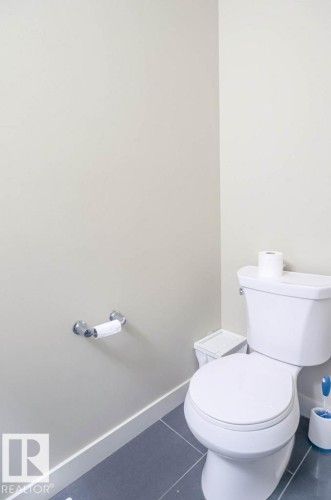 White toilet with a round bowl, chrome toilet paper holder, and dark gray tile flooring - 16619 14 Avenue, Edmonton, AB - Indoor Photo Showing Bathroom