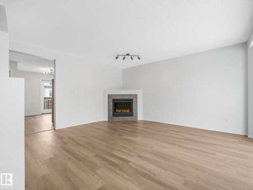 The living area features light-colored walls and flooring, with a corner fireplace showcasing a dark tile surround - 14 15128 22 Street, Edmonton, AB - Indoor Photo Showing Living Room With Fireplace