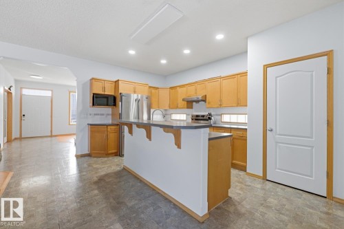 Kitchen featuring a central island with a bar-height counter, light wood cabinetry, stainless steel appliances, recessed ceiling lights, and patterned tile flooring - 9904 144 Avenue, Edmonton, AB - Indoor Photo Showing Kitchen