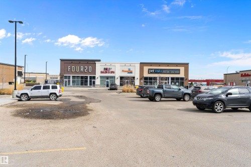 Commercial building featuring multiple retail storefronts, a prominent central entrance, varied exterior finishes including brick and paneling, large display windows, and an expansive paved parking lot - 3314 9 Street Nw, Edmonton, AB - Outdoor
