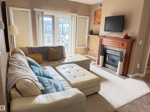 This inviting living space features a fireplace with a wood mantle, large windows with blinds, and a glass-paneled door - 438 1406 Hodgson Way, Edmonton, AB - Indoor Photo Showing Living Room With Fireplace