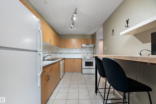 Kitchen featuring light wood cabinetry, white appliances, track lighting, and a breakfast bar with an integrated shelf - 206 9929 113 Street, Edmonton, AB - Indoor Photo Showing Kitchen With Double Sink