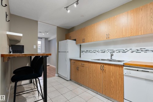 Kitchen featuring wood-finish cabinetry, a double basin stainless steel sink, a tile backsplash with decorative accents, and tiled flooring - 206 9929 113 Street, Edmonton, AB - Indoor Photo Showing Kitchen With Double Sink