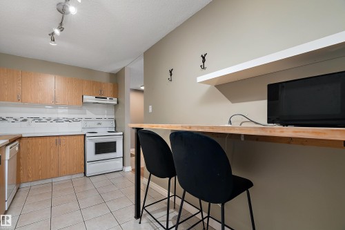 Kitchen featuring wood-finish cabinetry, tile flooring, white appliances, and a breakfast bar with an integrated floating shelf - 206 9929 113 Street, Edmonton, AB - Indoor Photo Showing Kitchen