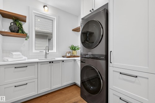 Dedicated utility space featuring a stacked washer and dryer, white shaker-style cabinetry with matte black hardware, wood-finish flooring, and a window above a stainless steel sink with a chrome faucet - 149 Eastgate Way, St. Albert, AB - Indoor Photo Showing Laundry Room