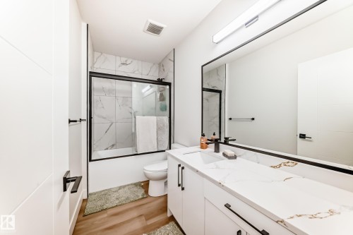 Contemporary bathroom featuring a spacious vanity with a white countertop and integrated sink, a rectangular mirror with a black frame, wood-finish flooring, a white bathtub with a sliding glass door, and tiled walls surrounding the tub - 17459 80 Street, Edmonton, AB - Indoor Photo Showing Bathroom