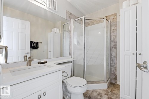 Bathroom featuring a corner shower enclosure, white vanity with integrated sink, a commode, a large wall-mounted mirror, and tile flooring - 115 10105 95 Street, Edmonton, AB - Indoor Photo Showing Bathroom
