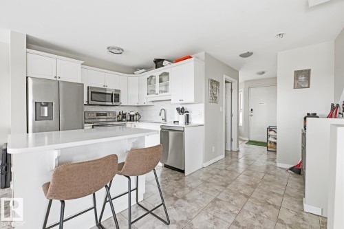 Modern kitchen featuring white cabinetry, stainless steel appliances, a white island with an integrated breakfast bar, and a patterned tile floor - 115 10105 95 Street, Edmonton, AB - Indoor Photo Showing Kitchen
