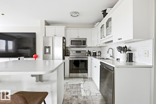 Modern kitchen featuring white cabinetry, stainless steel appliances, a white tile backsplash, and a light-toned countertop with an integrated breakfast bar - 115 10105 95 Street, Edmonton, AB - Indoor Photo Showing Kitchen