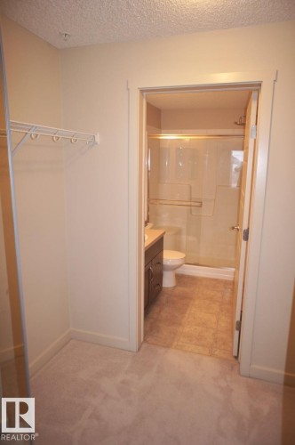 Carpeted dressing area with built-in wire shelving and direct access to a full bathroom featuring a vanity with storage, a toilet, a shower with a sliding glass door, and tiled flooring - 326 6076 Schonsee Way, Edmonton, AB - Indoor Photo Showing Bathroom