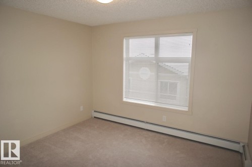 Carpeted room featuring a large window with horizontal blinds, neutral wall paint, and baseboard heating - 326 6076 Schonsee Way, Edmonton, AB - Indoor Photo Showing Other Room