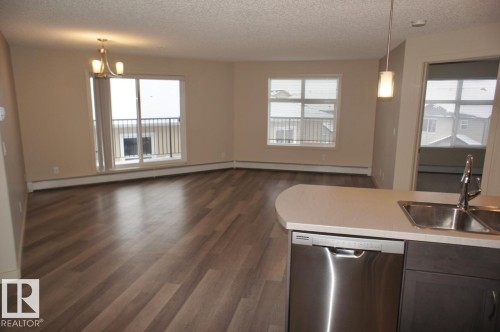 Open-concept living area with wood-finish flooring, a sliding glass door leading to a balcony with black railings, and a large window providing ample natural light - 326 6076 Schonsee Way, Edmonton, AB - Indoor Photo Showing Kitchen With Double Sink