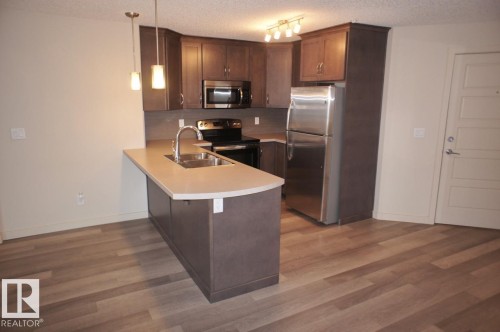 Kitchen featuring wood-finish flooring, dark wood cabinetry, a stainless steel refrigerator, and a kitchen island with an integrated sink and pendant lighting - 326 6076 Schonsee Way, Edmonton, AB - Indoor Photo Showing Kitchen With Double Sink