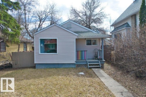 Single-story residence featuring light-colored horizontal siding, a covered front porch with lattice detailing, a shingled roof, a paved walkway, and a front yard area - 12207 110 Avenue, Edmonton, AB - Outdoor