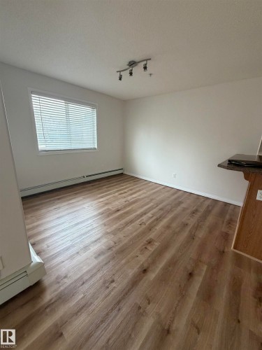 Open-concept room featuring wood-finish flooring, white walls, and a large window with horizontal blinds - 104 8119 112 Avenue Nw, Edmonton, AB - Indoor Photo Showing Other Room