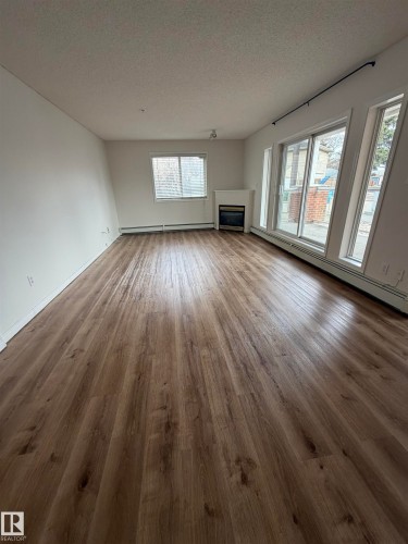 Spacious room featuring wood-finish flooring, a built-in fireplace with a white mantle, a large window with blinds, and a sliding glass door flanked by two vertical windows - 104 8119 112 Avenue Nw, Edmonton, AB - Indoor Photo Showing Other Room With Fireplace