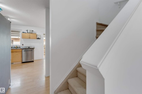 Carpeted staircase with white handrail and partial view of a kitchen featuring a stainless steel dishwasher, wood-finish cabinetry, and light wood-finish flooring - 48 2503 24 Street, Edmonton, AB - Indoor