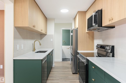 Galley kitchen featuring light wood-finish upper cabinetry, dark green lower cabinetry, and stainless steel appliances - 15C Callingwood Crest, Edmonton, AB - Indoor Photo Showing Kitchen With Upgraded Kitchen