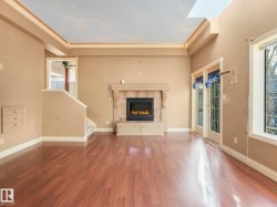 Spacious room featuring wood-finish flooring, a stone fireplace with built-in cabinetry, dual French doors, a large picture window, and a tray ceiling with a skylight - 