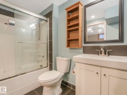 Bathroom featuring a shower/tub combination with sliding glass doors, a white vanity with a rectangular basin, a framed mirror, a wall-mounted wooden shelving unit, and dark floor tiles - 