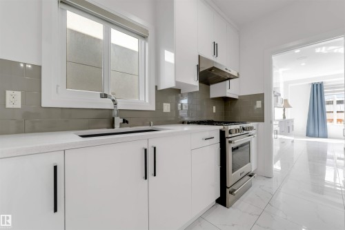Contemporary kitchen featuring white cabinetry with black hardware, stainless steel gas range with coordinating hood, integrated sink with chrome faucet, light-toned countertops, and a gray subway tile backsplash - 1923 19 Avenue, Edmonton, AB - Indoor Photo Showing Kitchen With Upgraded Kitchen