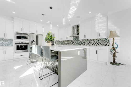 Modern kitchen featuring an expansive center island with seating, white shaker-style cabinetry, a mosaic tile backsplash, and integrated stainless steel appliances - 1923 19 Avenue, Edmonton, AB - Indoor Photo Showing Kitchen With Upgraded Kitchen