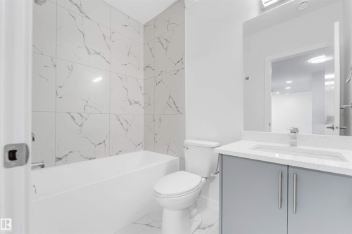 Bathroom featuring a white bathtub with marble-look tile surround, a built-in toilet, a single-sink vanity with a light gray cabinet base, and a chrome faucet - 1923 19 Avenue, Edmonton, AB - Indoor Photo Showing Bathroom