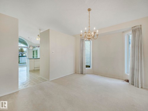 Spacious room featuring neutral carpeting, a brass chandelier, two vertical windows with blinds and drapes, and an open entryway to a tiled area - 23 9731 174 Street, Edmonton, AB - Indoor Photo Showing Other Room