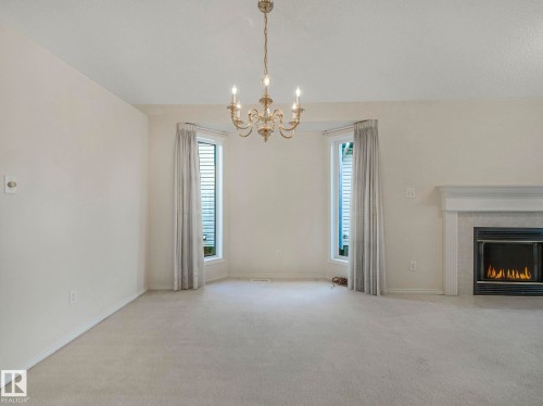 Spacious room featuring a central chandelier, two bay windows with vertical blinds, a fireplace with a light-colored mantel, and neutral carpeting - 23 9731 174 Street, Edmonton, AB - Indoor With Fireplace
