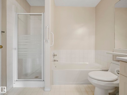 Bathroom featuring a walk-in shower with frosted glass door, a bathtub with grab bars, a toilet, and light-toned wall and floor tiles - 23 9731 174 Street, Edmonton, AB - Indoor Photo Showing Bathroom