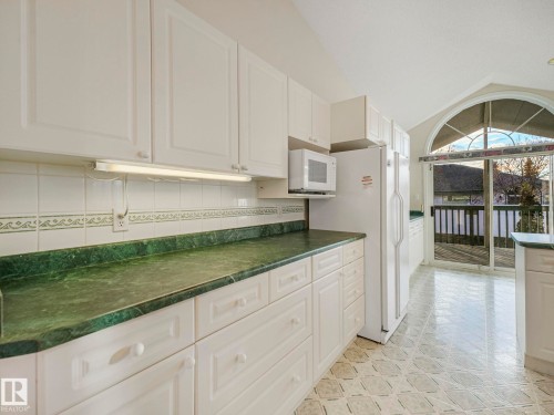 Kitchen featuring white cabinetry, green stone countertops, white tile backsplash with decorative accent, patterned floor tile, and a vaulted ceiling with an arched window above sliding glass doors - 23 9731 174 Street, Edmonton, AB - Indoor Photo Showing Other Room