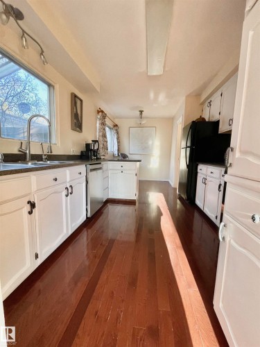 Kitchen featuring rich wood-finish flooring, white cabinetry with dark hardware, stainless-steel sink with gooseneck faucet, black refrigerator, and a recessed ceiling light fixture - 11681 72 Avenue, Edmonton, AB - Indoor Photo Showing Kitchen
