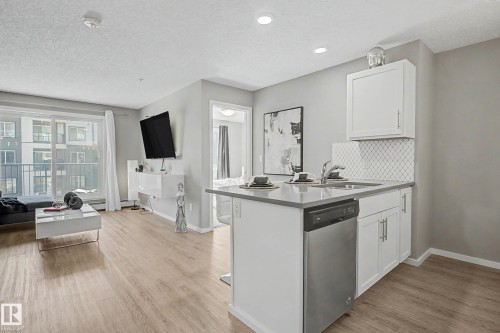 Open-concept living space featuring wood-finish flooring, a kitchen island with integrated dishwasher, white shaker cabinetry, and a diamond-patterned tile backsplash - 217 394 Windermere Road, Edmonton, AB - Indoor Photo Showing Kitchen