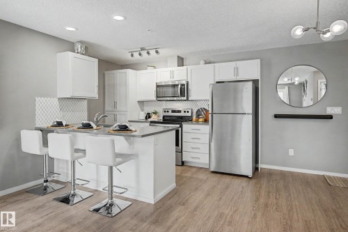Modern kitchen featuring white cabinetry, stainless steel appliances, a white diamond-patterned tile backsplash, a breakfast bar, and wood-finish flooring - 217 394 Windermere Road, Edmonton, AB - Indoor Photo Showing Kitchen With Stainless Steel Kitchen
