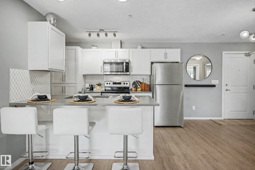 Kitchen featuring white cabinetry, stainless steel appliances, a breakfast bar, light wood-finish flooring, and a patterned tile backsplash - 217 394 Windermere Road, Edmonton, AB - Indoor Photo Showing Kitchen With Stainless Steel Kitchen With Double Sink With Upgraded Kitchen