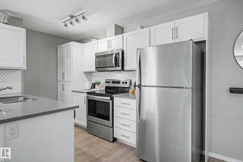 Modern kitchen featuring white shaker cabinetry, stainless steel appliances, a diamond-patterned tile backsplash, grey countertops, and wood-finish flooring - 217 394 Windermere Road, Edmonton, AB - Indoor Photo Showing Kitchen With Stainless Steel Kitchen With Double Sink