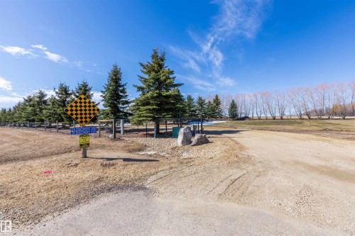 Gravel driveway entrance with a natural stone accent, bordered by a row of mature evergreen trees and a wooden fence - 2B Discovery Avenue, Rural Sturgeon County, AB 