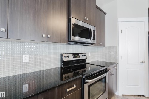 Contemporary kitchen featuring dark wood-finish cabinetry with modern hardware, stainless steel microwave, black glass-top electric range, and a mosaic tile backsplash - 611 176 Street, Edmonton, AB - Indoor Photo Showing Kitchen