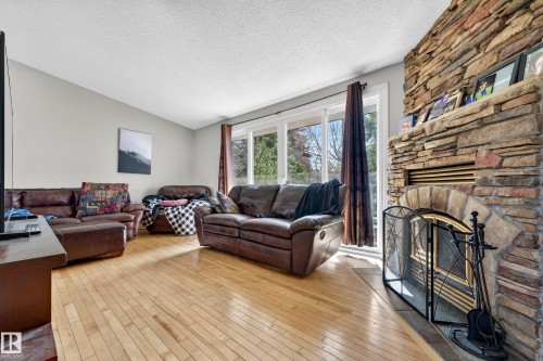 Living area featuring a floor-to-ceiling natural stone fireplace, light wood-finish flooring, and expansive glass patio doors - 1 Greenwich Crescent, St. Albert, AB - Indoor Photo Showing Living Room With Fireplace