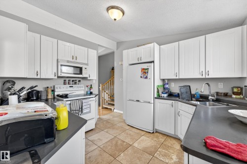 Kitchen featuring white cabinetry, dark countertops, a stainless steel sink with a gooseneck faucet, white appliances, and tan tile flooring - 1 Greenwich Crescent, St. Albert, AB - Indoor Photo Showing Kitchen With Double Sink